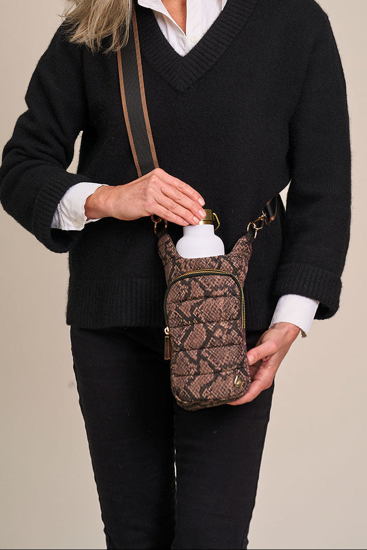 A woman wearing a black sweater and white collared shirt models a brown and black snakeskin-patterned crossbody water bottle bag, shown with a white water bottle inserted.